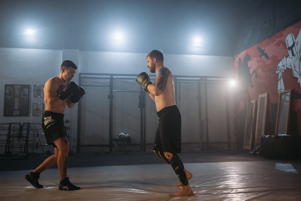 services-04 Two men sparring in a boxing gym, showcasing focus and determination under dramatic lighting.