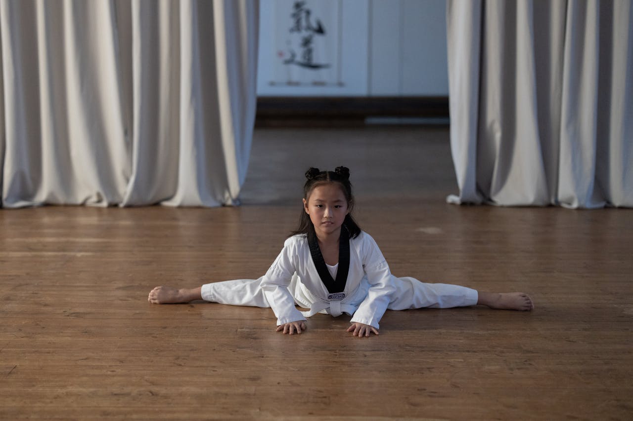 services-02 A young girl in a dobok performing a split on a wooden floor during a taekwondo practice indoors.