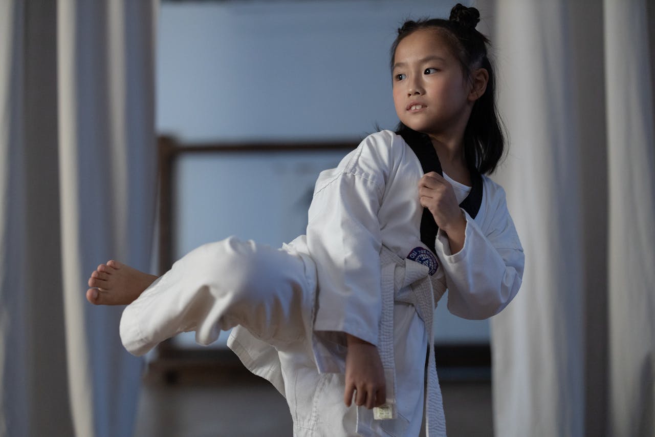 services-04 A young girl in a dobok practicing a taekwondo kick indoors, showcasing martial arts skills.
