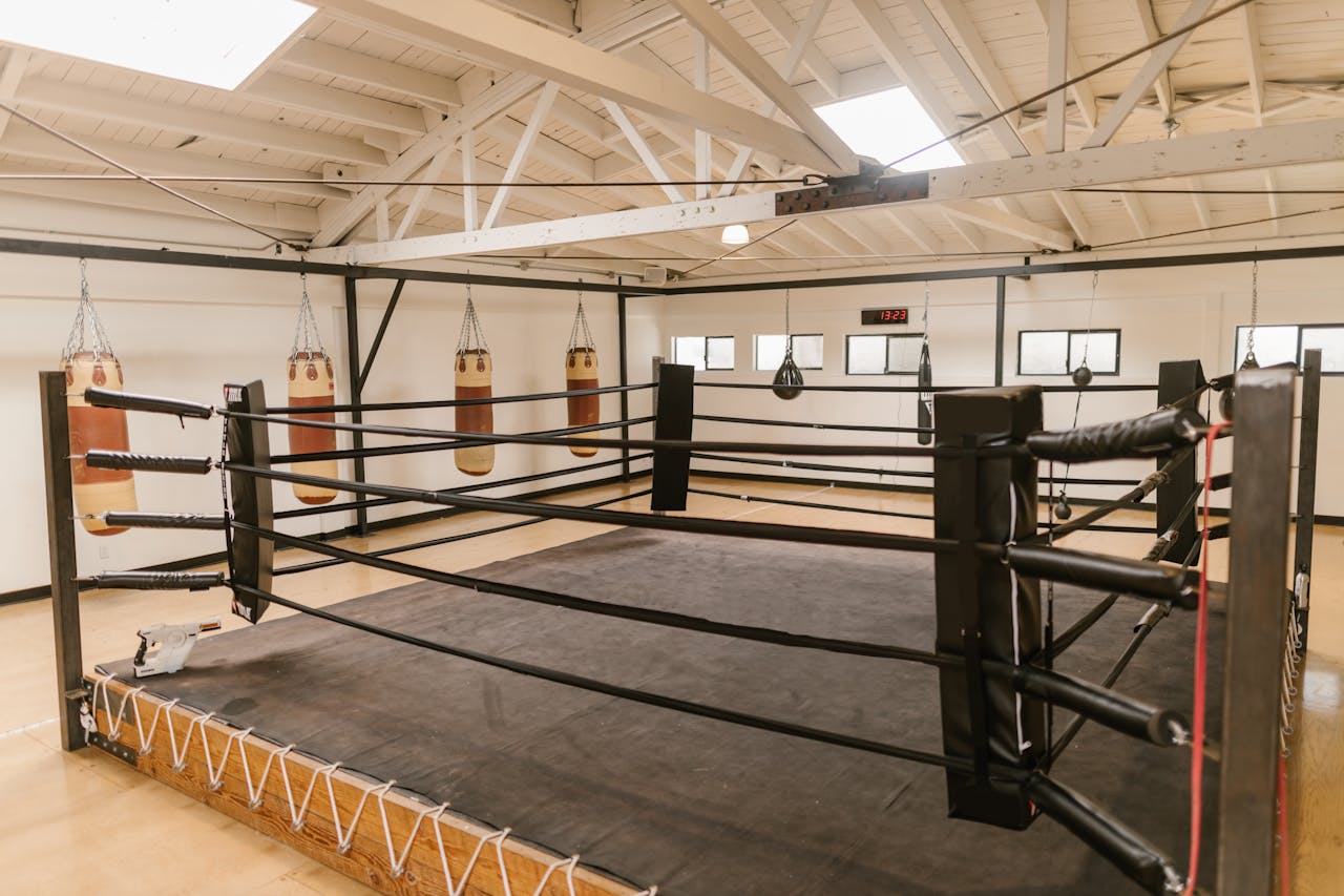 services-03 Interior view of a spacious boxing gym featuring a central ring and multiple heavy punching bags.